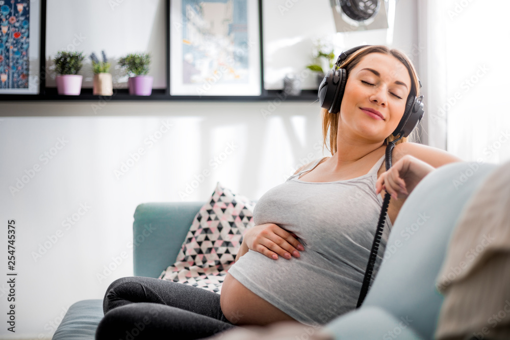 Relaxed pregnant woman sitting on sofa at home and listening music in headphones