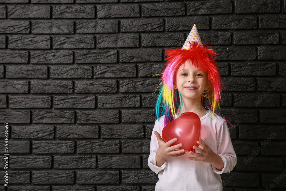 Funny little girl in bright wig and with balloon on dark background. April fools day celebration