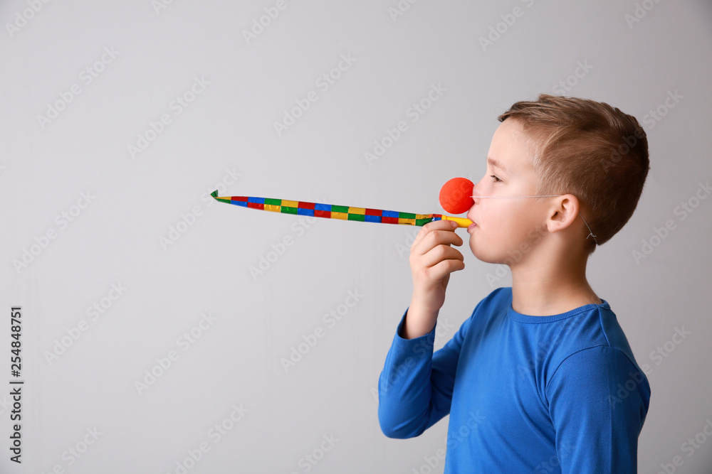 Funny little boy with clown nose and party whistle on light background. April fools day celebration