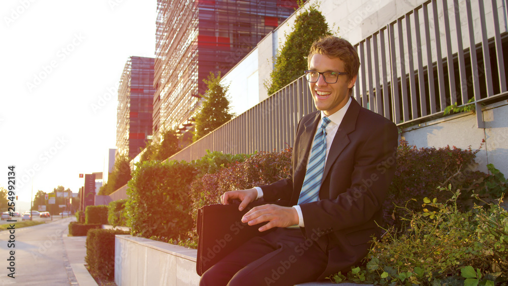 LENS FLARE: Cheerful businessman sitting on a ledge and singing after promotion