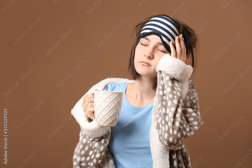 Sleepy young woman with mask and cup of coffee on color background