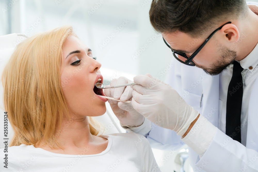 Young handsome dentist examining teeth of happy woman patient sitting on dentist chair in dental cli