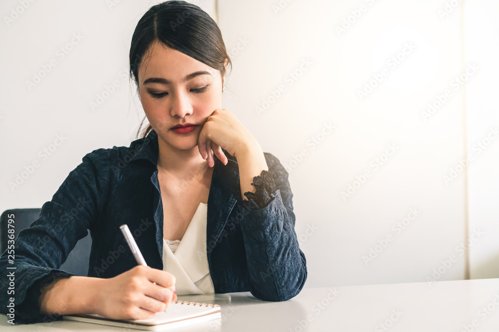 Beautiful young Asian woman writes letter on notebook while sitting at office desk. Content writer a