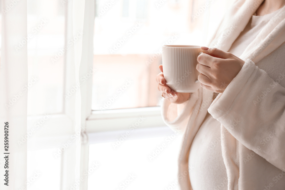 Pregnant woman with cup of hot tea standing near window at home