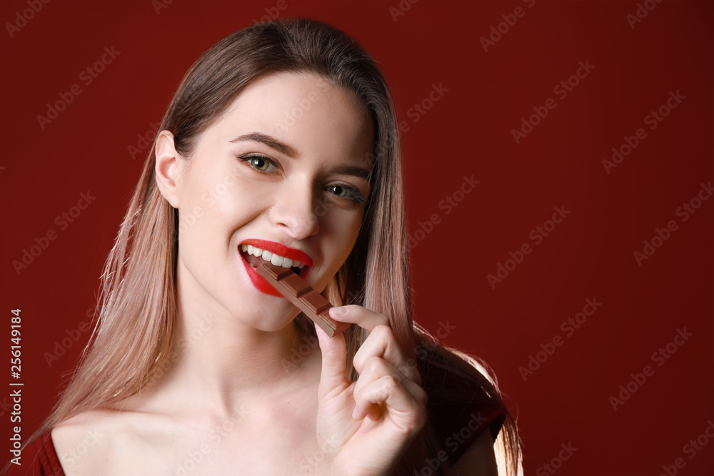 Beautiful young woman eating tasty chocolate on color background