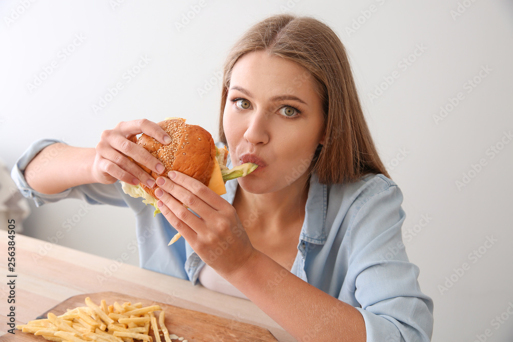 Young woman eating tasty burger with french fries at table