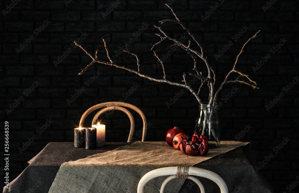 Pomegranates with tree branches in jar and candles on table against dark background