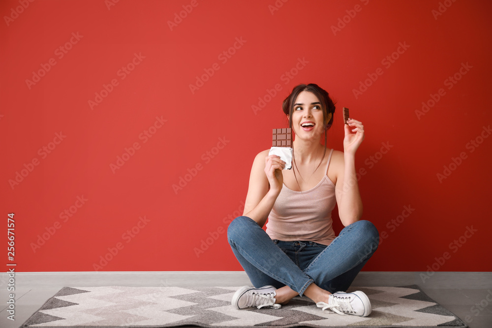 Beautiful young woman with tasty chocolate sitting on floor near color wall