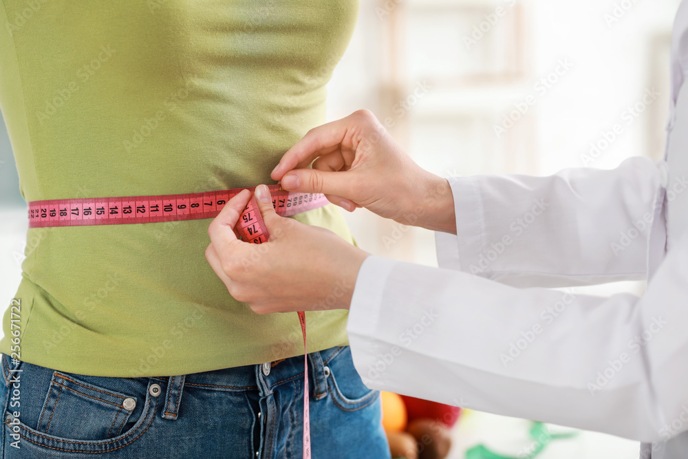 Nutritionist measuring waist of young woman in weight loss clinic, closeup