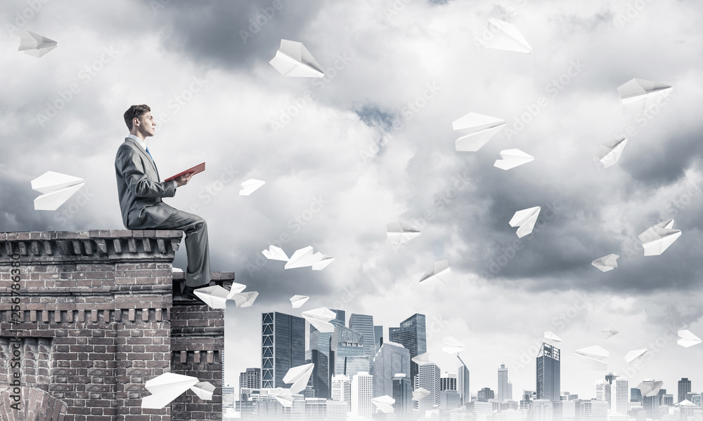 Man on roof edge reading book and cityscape at background