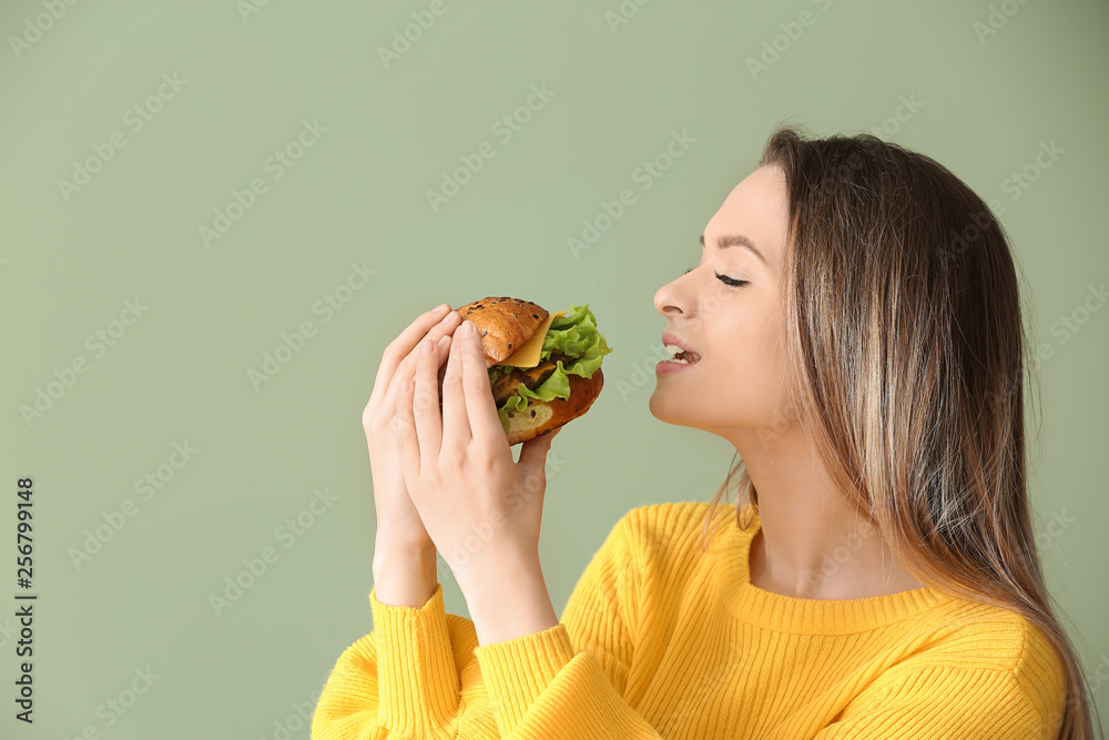 Beautiful young woman eating tasty burger on color  background