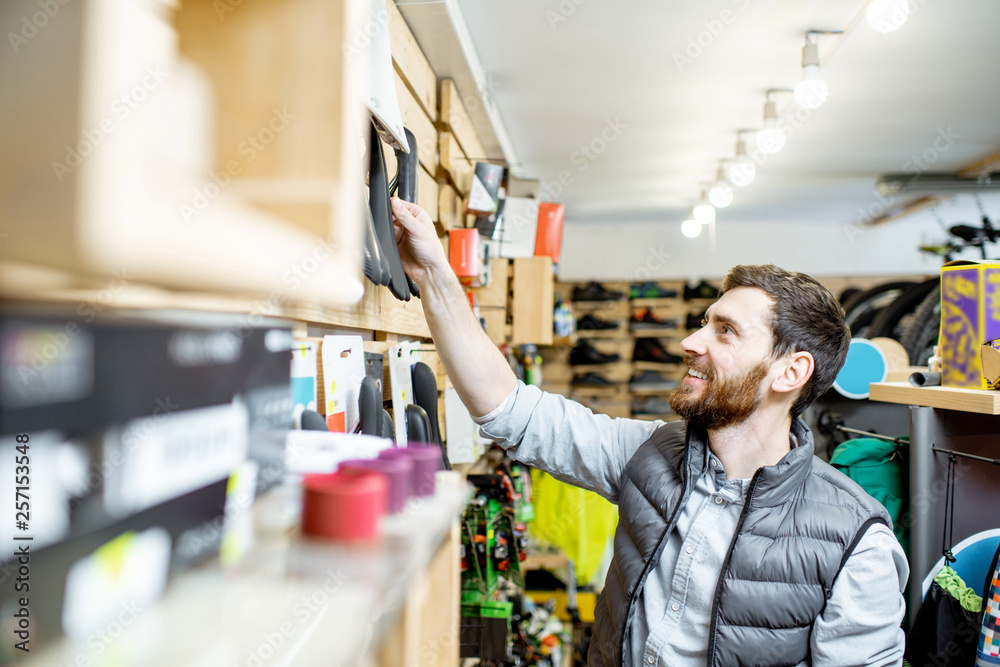 Man choosing a bicycle saddle standing near the shelves with bicycle parts in the sports shop