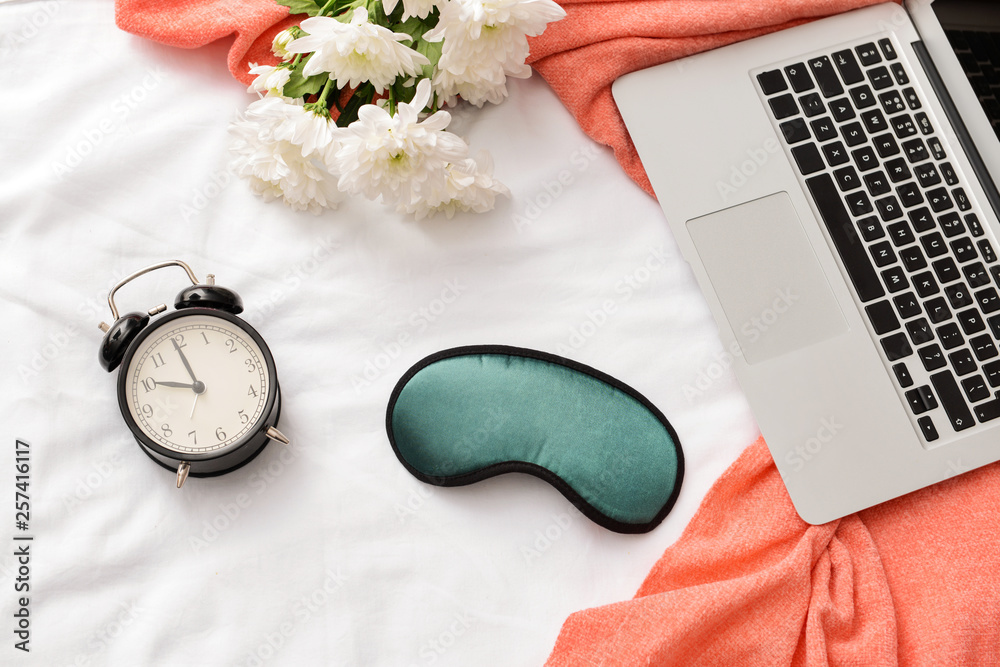 Composition with sleep mask, laptop, clock and flowers on bed