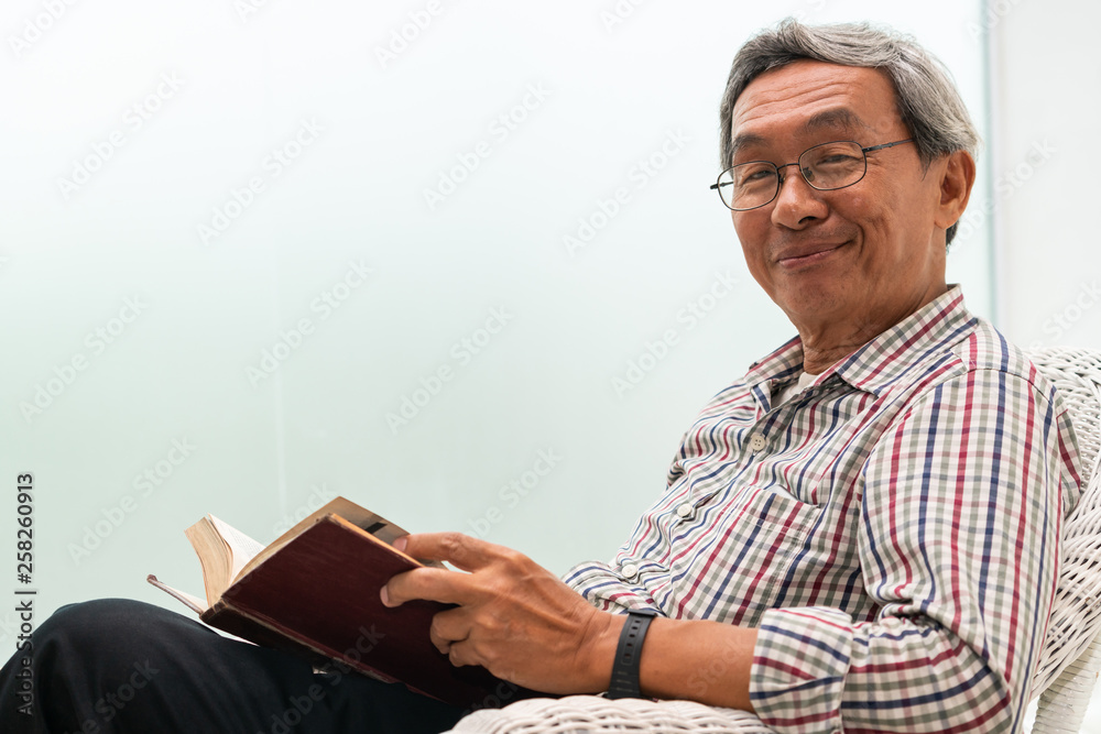 Happy senior Asian man reading book on the chair in living room at home. Retirement lifestyle and ag