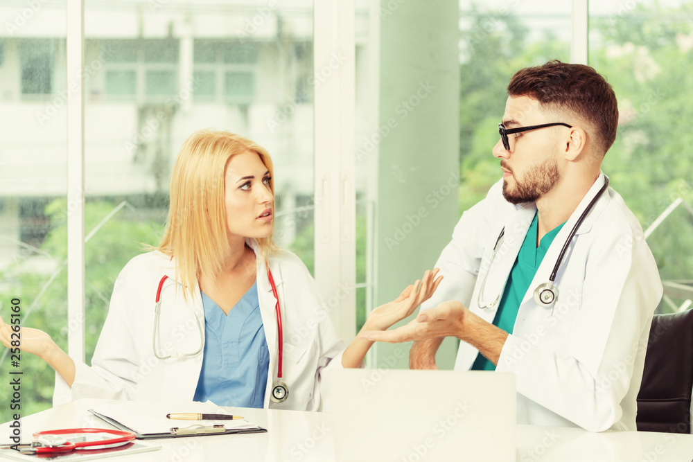 Young female doctor works at office in hospital while talking to male doctor sitting beside her. Med