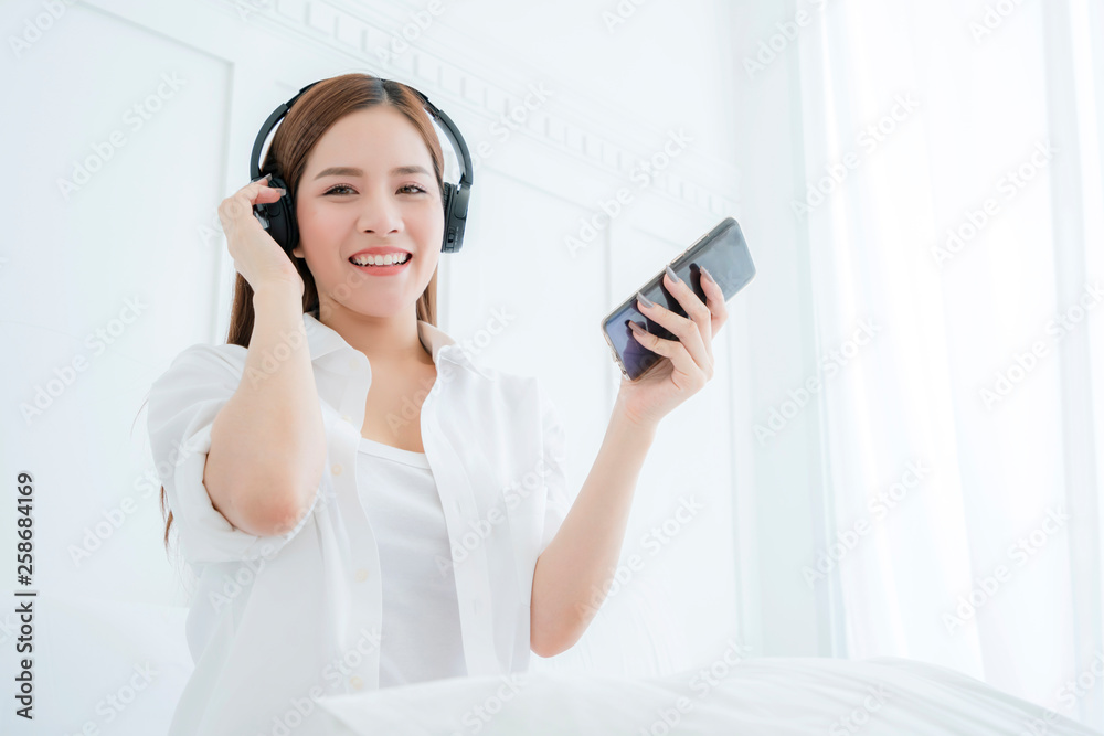 beautiful attractive asian woman listen song from headphone hand hold smartphone white bedroom backg