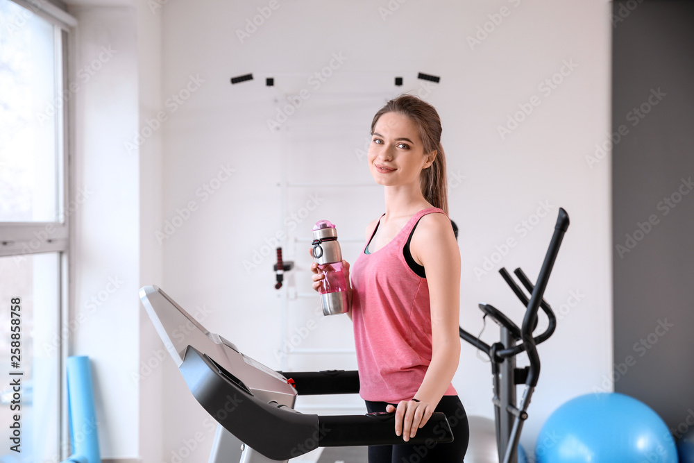 Sporty young woman drinking water after training on treadmill in gym