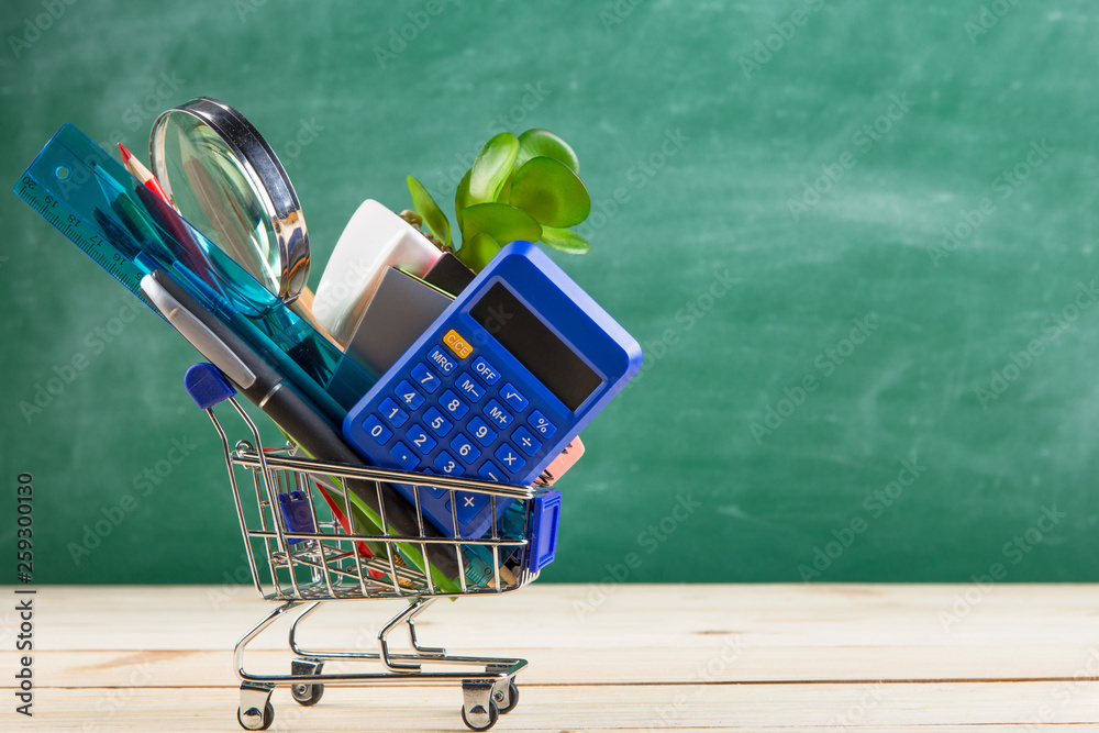 Education concept - school supplies in a shopping cart on the desk in the auditorium, blackboard bac