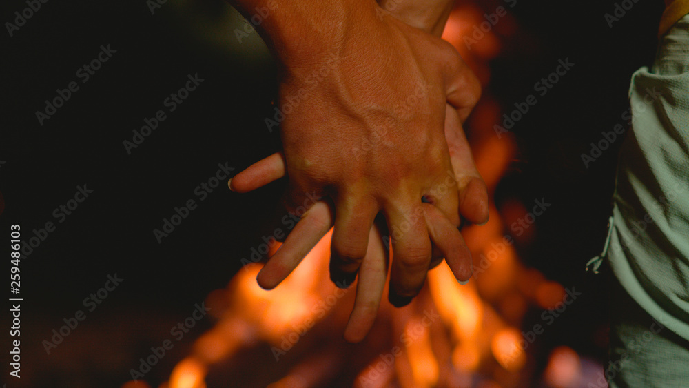 MACRO: Unrecognizable young man and woman holding hands over the campfire.