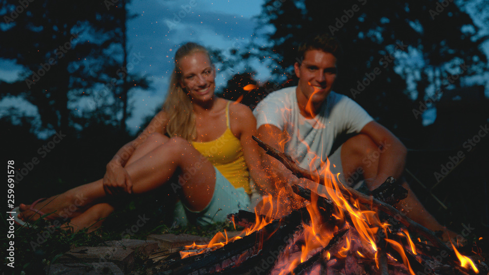 CLOSE UP: Cheerful man pokes fire with a stick while camping with girlfriend.