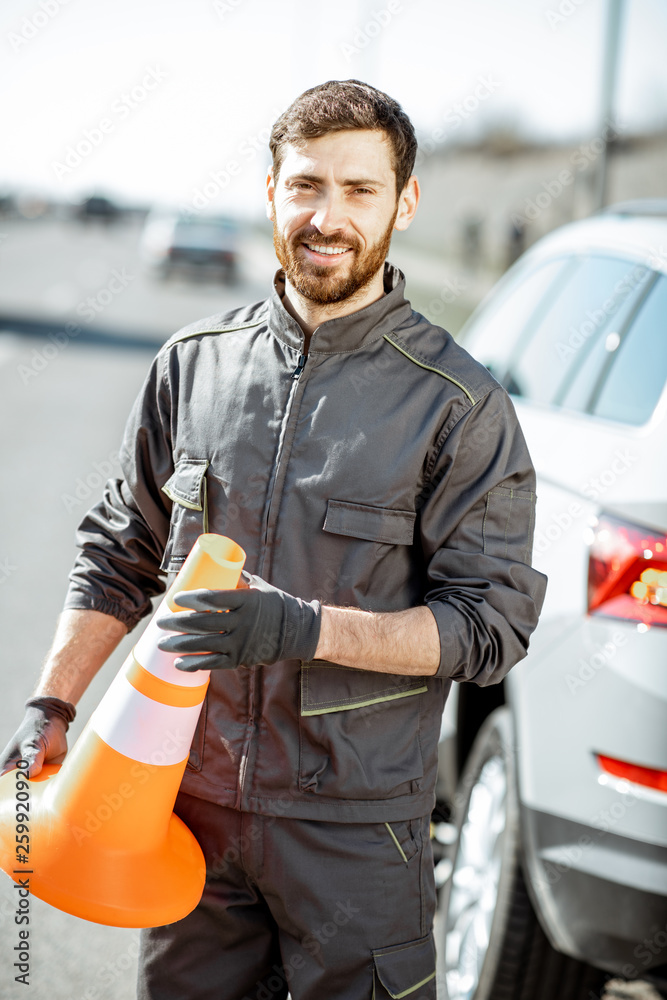 Portrait of a happy road assistance worker in uniform standing with cone near the car on the roadsid