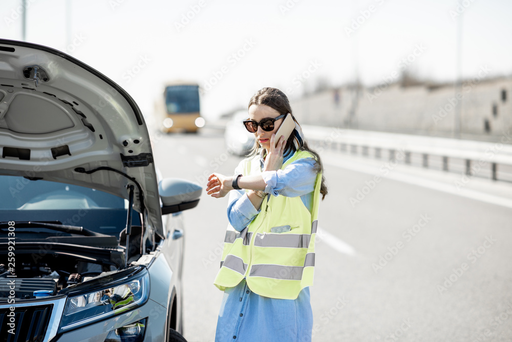 一名尴尬的女子站在高速公路路边的一辆坏汽车附近呼叫道路救援