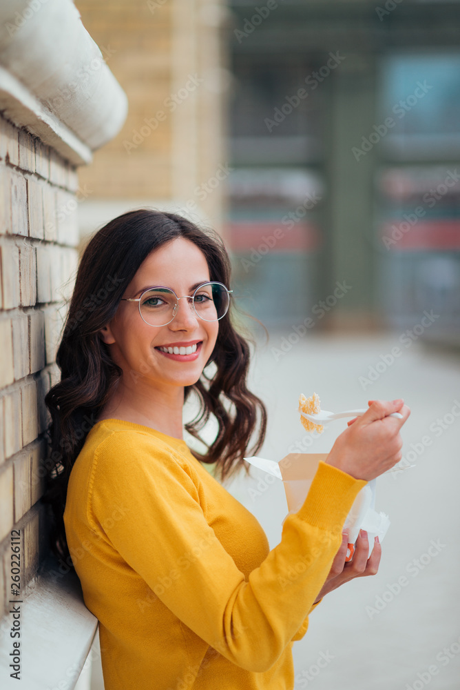 Portrait of a young woman eating from takeaway box outdoors, leaning on a wall and smiling at camera