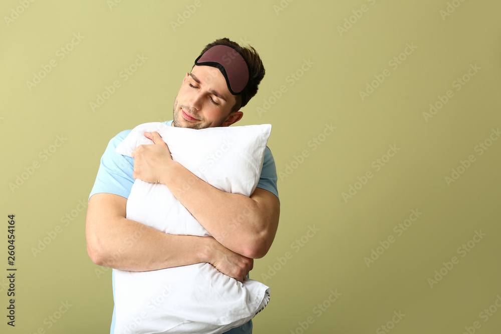 Young man with sleep mask and pillow on color background