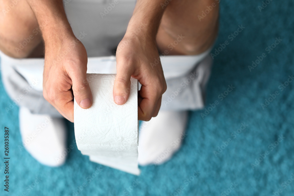 Man with roll of paper sitting on toilet bowl at home, closeup