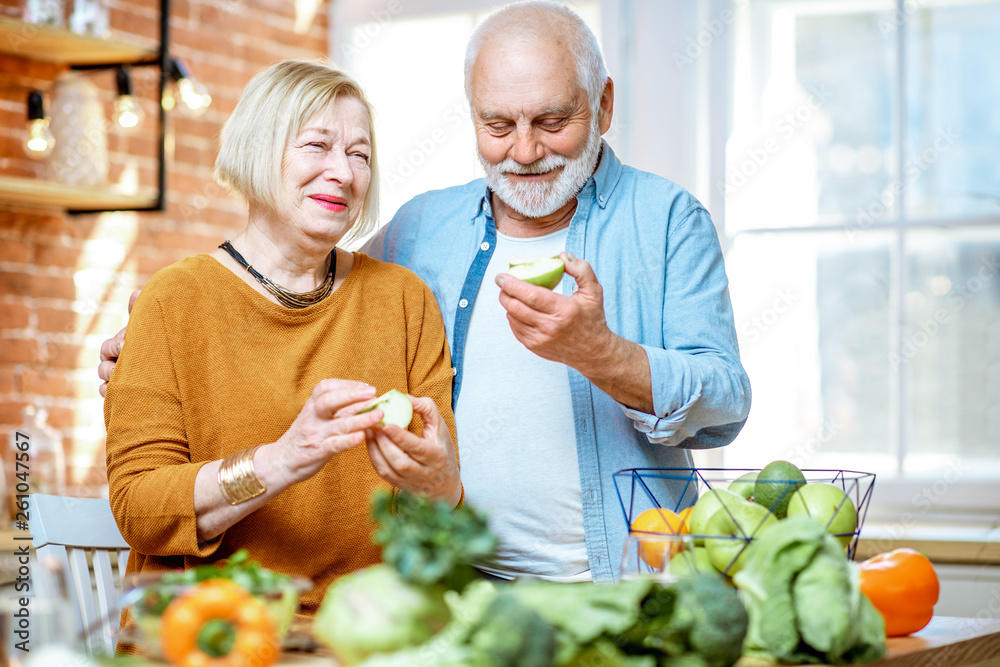 Portrait of a cheerful senior couple eating apples standing together with healthy food on the kitche