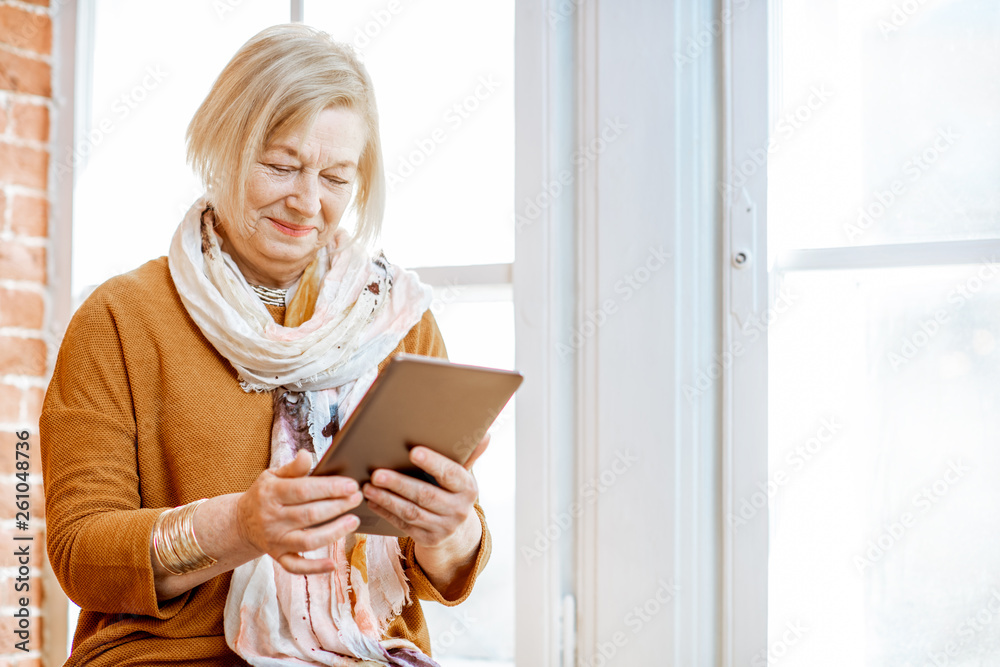 Beautiful senior woman using digital tablet sitting near the window at home