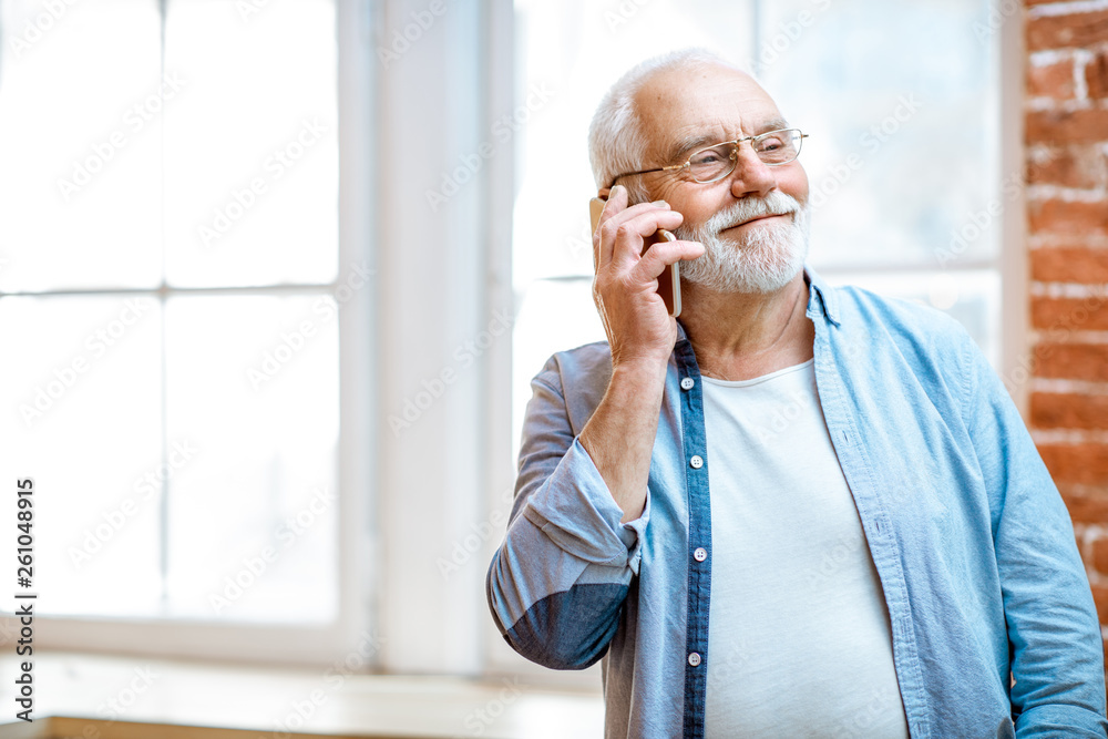 Cheerful senior grandfather talking with phone near the window at home