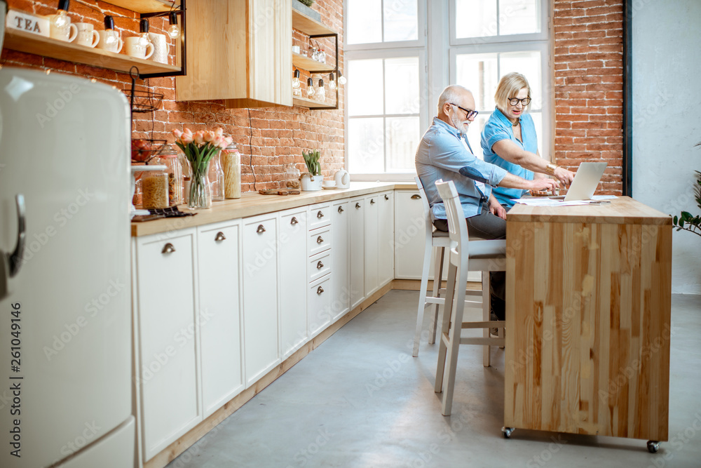 Senior couple sitting together with laptop on the kitchen at home. Wide interior view