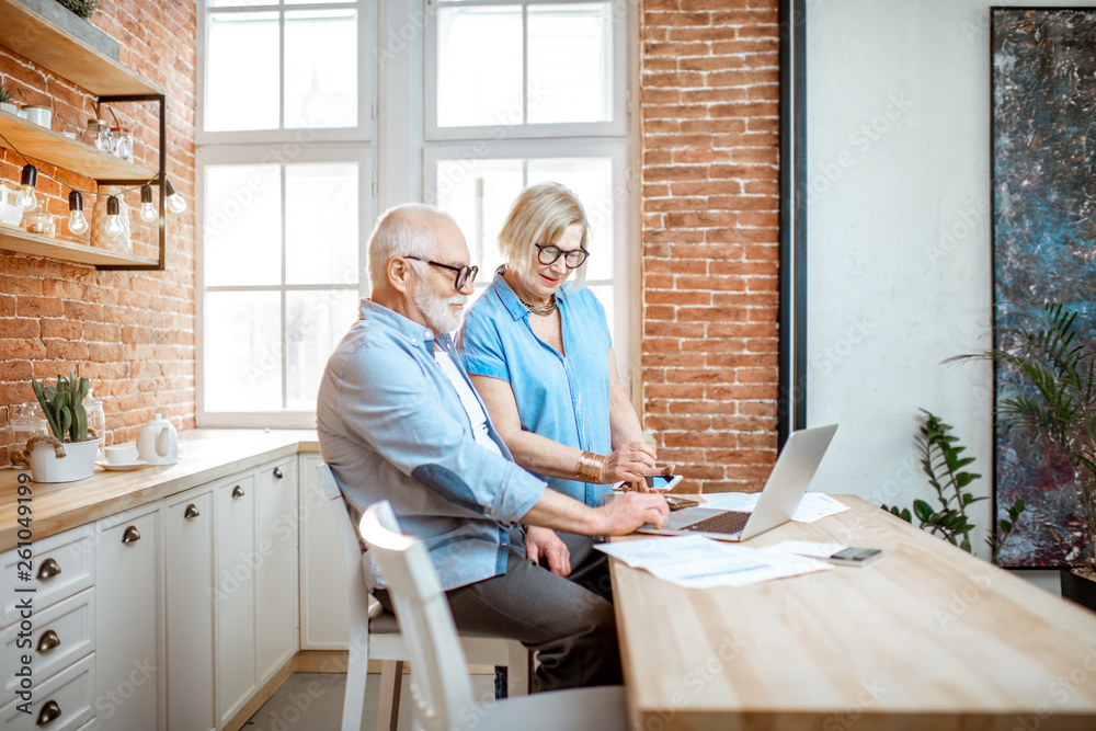 Beautiful senior couple in blue shirts sitting together with laptop on the kitchen at home