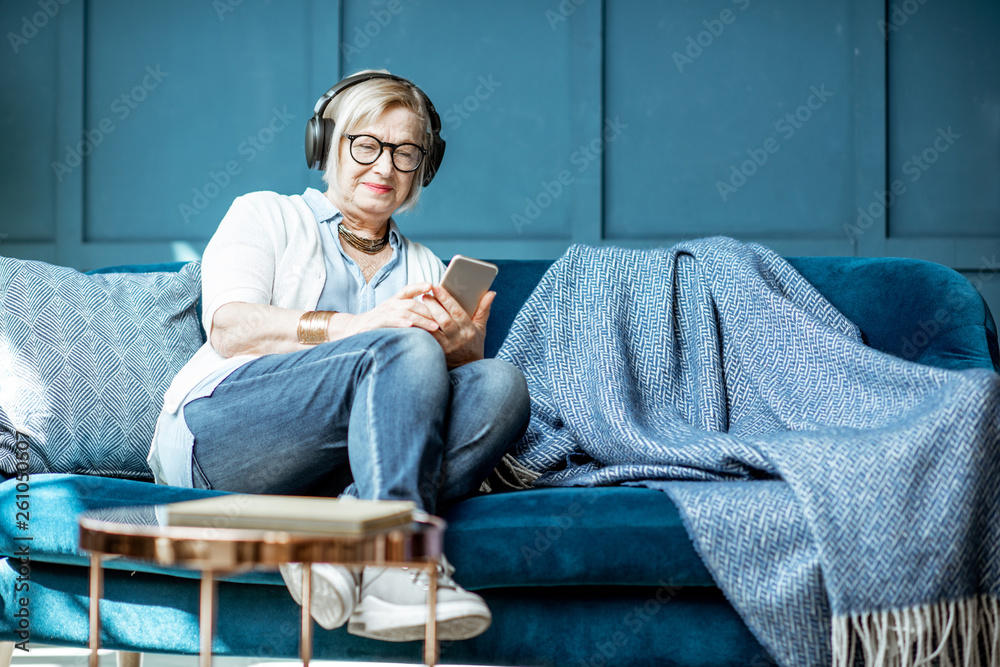 Senior woman dressed casually listening to the music with headphones and phone on the couch at home