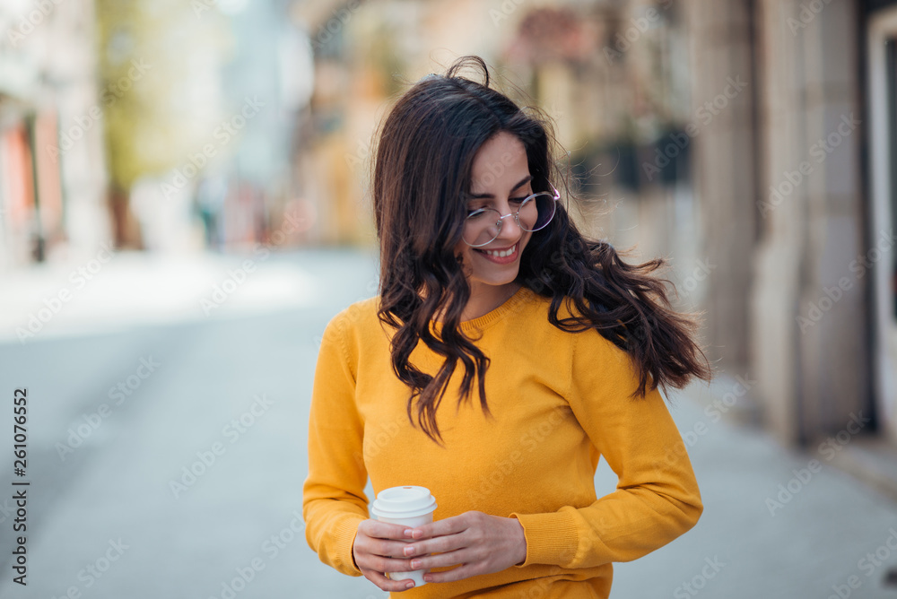 Cheerfull millenial woman walking in the city with takeaway coffee.