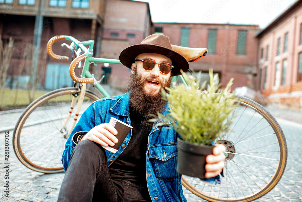 Portrait of a stylish hipster with flower pot sitting near his retro bicycle on the street on the ur