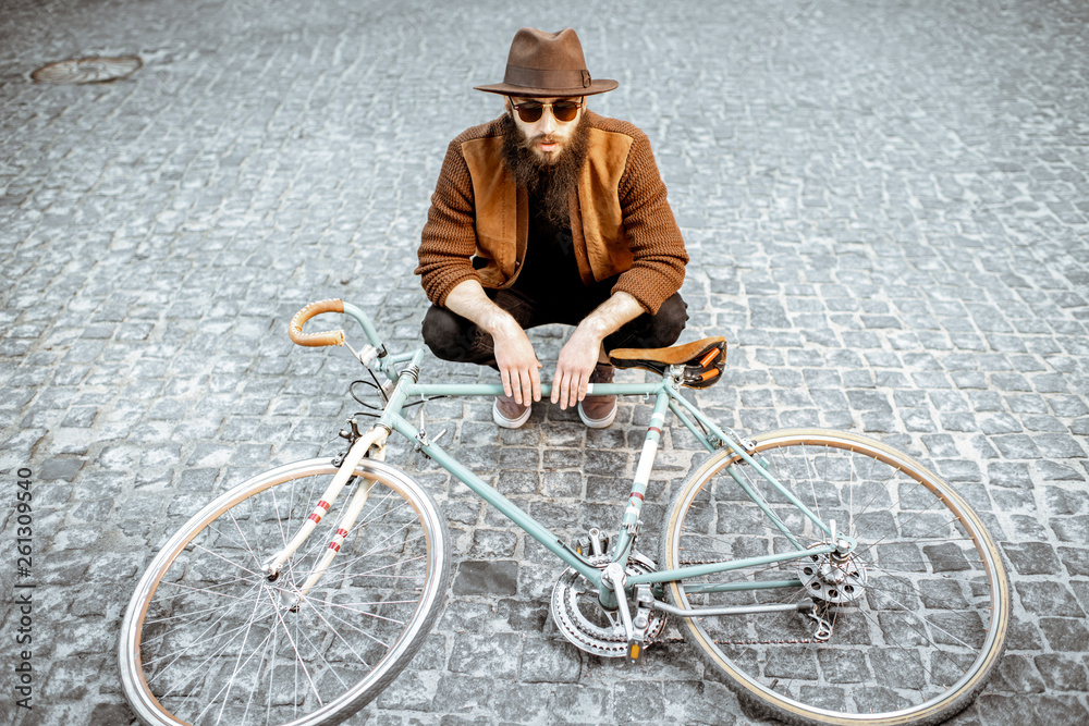 Portrait of a bearded hipster dressed stylishly with brown hat and sweater sitting with retro bicycl