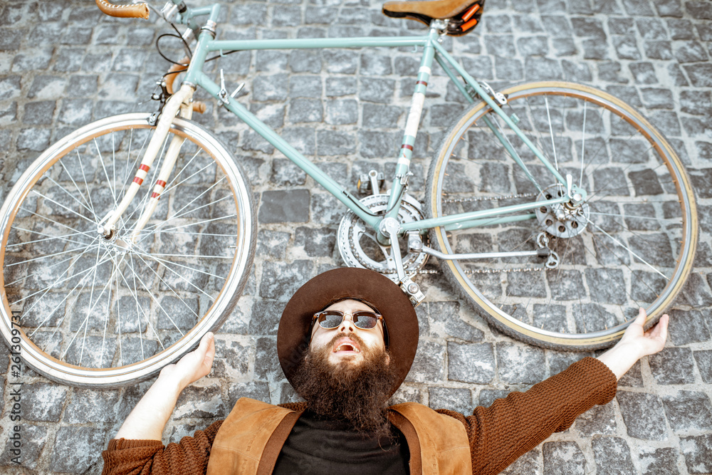 Portrait of a stylish bearded man in hat lying with retro bicycle on the pavement street, view from 