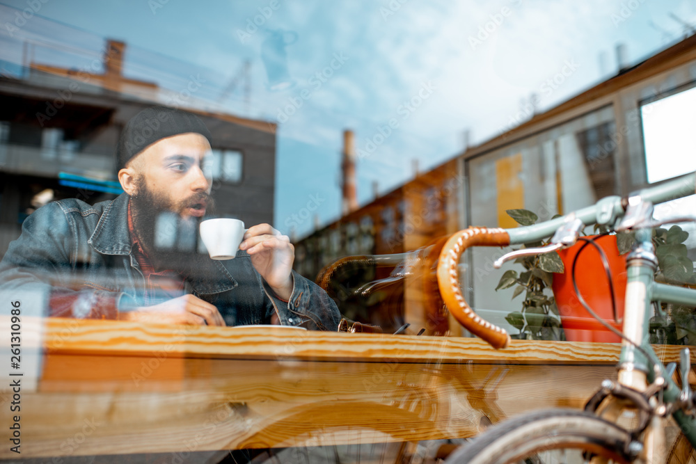 Stylish man enjoying a coffee drink while sitting at the cafe near the window with retro bicycle. Vi