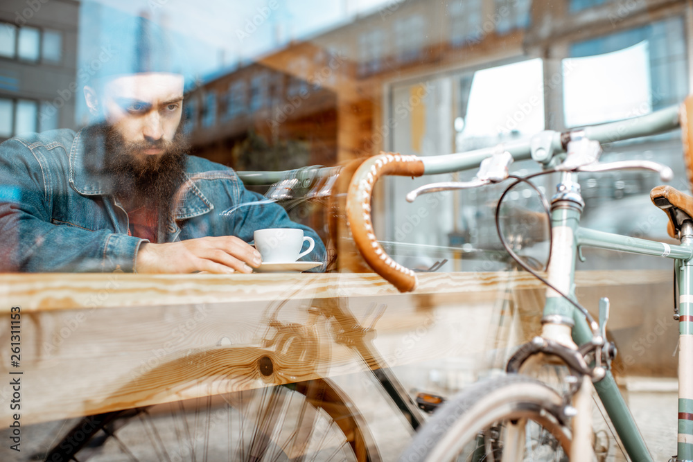 Stylish man enjoying a coffee drink while sitting at the cafe near the window with retro bicycle. Vi