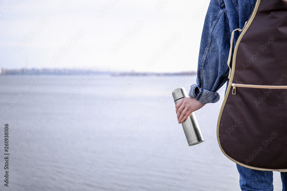 Woman with thermos and guitar near river, back view