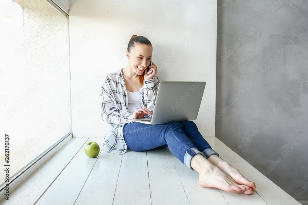 Young woman talking by phone while working on laptop indoors