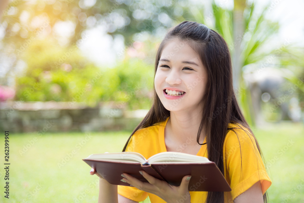 Pretty asian female teenager smiling while reading book in park