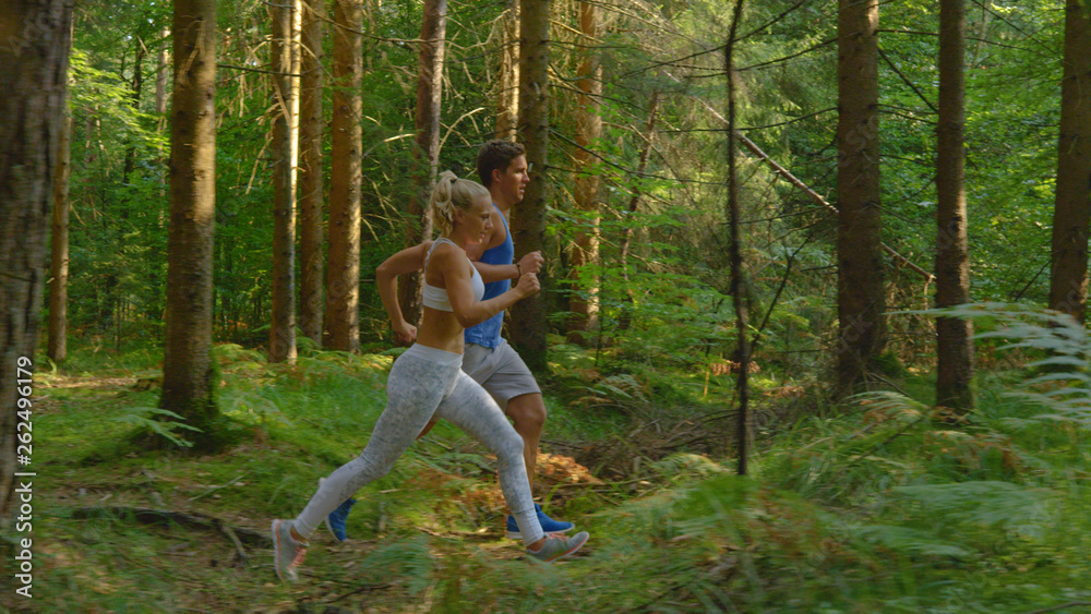 Athletic Caucasian man and his sporty girlfriend jog along sunlit forest trail.