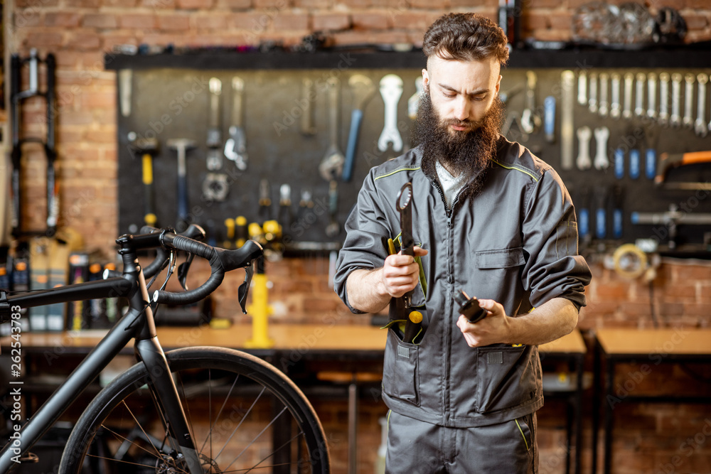 Portrait of a handsome bearded repairman in workwear standing with wrenches at the bicycle workshop