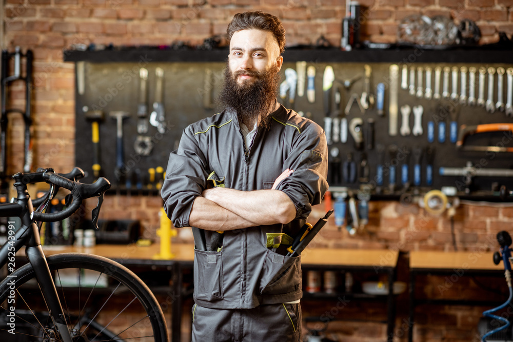 Portrait of a handsome bearded repairman in workwear standing with wrenches at the bicycle workshop