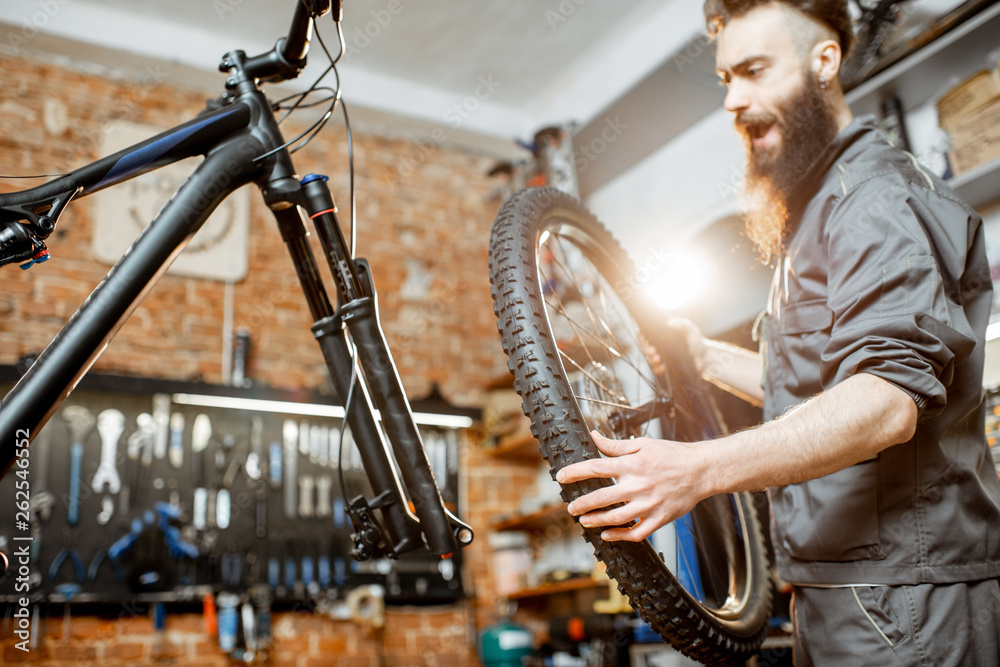 Handsome repairman in workwear serving mountain bicycle, standing with front wheel at the workshop o