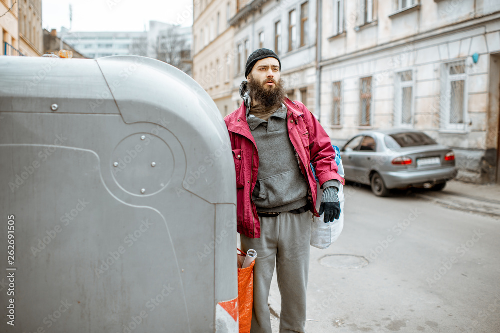 Portrait of a homeless depressed beggar standing with bags near the trash containers in the city. Co