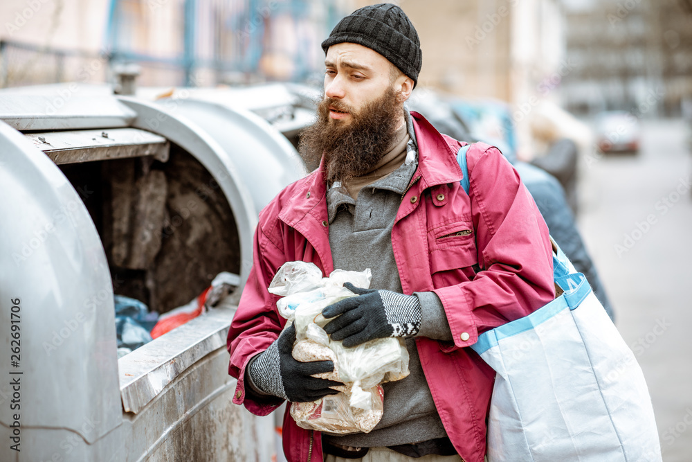 Portrait of a homeless bearded beggar standing with bag and some finded food near the trash containe